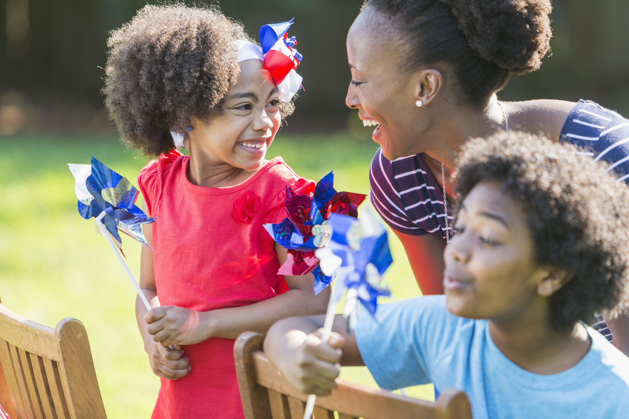 An African American mother with two mixed race children celebrating the Fourth ofJuly, an American patriotic holiday. They are playing with red, white and blue pinwheels, smiling and laughing, outdoors on a bright, sunny day. 